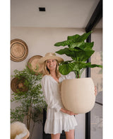 Une femme vêtue d'une robe blanche et d'un chapeau sourit tout en tenant la plante artificielle Calathea UV 72 cm à l'intérieur, avec des paniers tressés et une autre plante en arrière-plan.