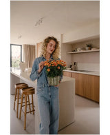 Une femme aux cheveux blonds bouclés, vêtue d'une chemise en jean et d'un jean, se tient dans une cuisine moderne, tenant une plante UV artificielle marguerite de 31 cm avec des fleurs orange vif. À côté d’elle se trouvent deux tabourets en bois près de l’îlot de cuisine.