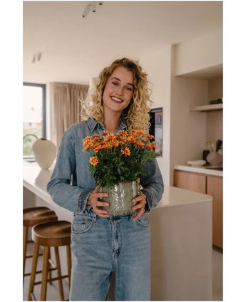 Une femme aux cheveux blonds bouclés, vêtue de vêtements en jean, sourit en tenant la plante Artificielle Daisy UV 31 cm – un pot de fleurs orange vif et jaune – dans une cuisine moderne.