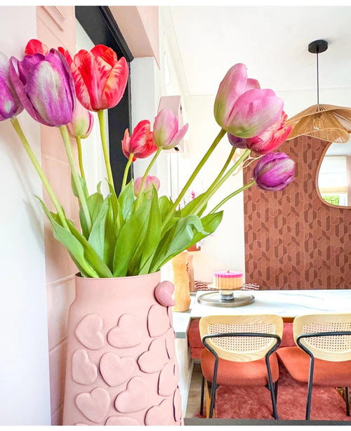 Un vase rose avec des cœurs en relief contient la fleur artificielle French Tulip 60 cm rouge sur une table à manger. Derrière, se trouvent des chaises avec dossier en osier, un gâteau sur la table et un miroir sur un mur marron à motifs sous une suspension tressée.
