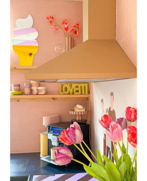 Une cuisine rose confortable a une tulipe française artificielle de 60 cm rouge au premier plan, des tasses de couleur pastel sur une étagère, une assiette LOVETTI, une peinture murale de deux personnes et un miroir fantaisiste en forme de cornet de glace au mur.