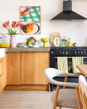 Une cuisine lumineuse avec des armoires en bois, une cuisinière noire, des serviettes vertes, de la vaisselle colorée et une photo de pâtisserie encadrée sur une table à carreaux prend un nouveau look grâce à la Fleur Artificielle Perruche Française Tulipe 76 cm rouge/jaune. Une chaise blanche confortable se trouve à côté d’une table à manger en bois clair.