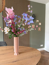 Un grand vase Ă rayures roses et blanches avec le bouquet de fleurs artificielles en soie Amethyst Romance se trouve sur une table ronde en bois dans une piĂšce moderne avec des murs verts, des boiseries et de petites Ćuvres d'art encadrĂ©es.