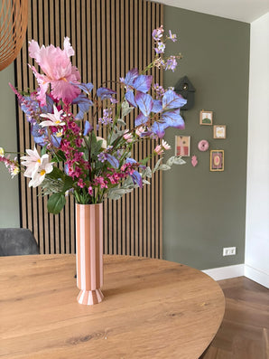 Un grand vase à rayures roses et blanches avec le bouquet de fleurs artificielles en soie Amethyst Romance se trouve sur une table ronde en bois dans une pièce moderne avec des murs verts, des boiseries et de petites œuvres d'art encadrées.