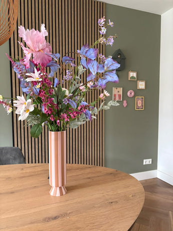 Un grand vase Ă rayures roses et blanches avec le bouquet de fleurs artificielles en soie Amethyst Romance se trouve sur une table ronde en bois dans une piĂšce moderne avec des murs verts, des boiseries et de petites Ćuvres d'art encadrĂ©es.
