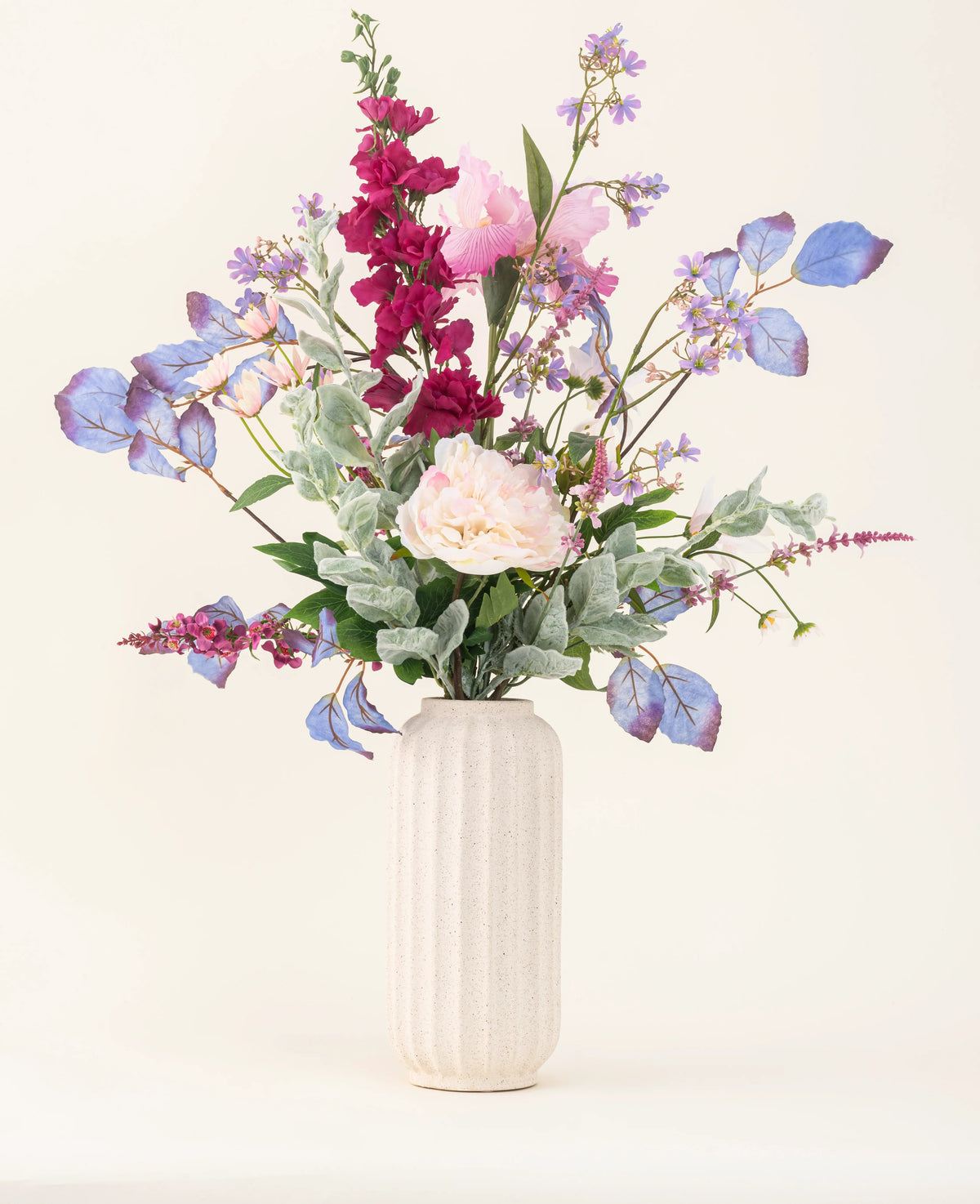 Un vase blanc texturé présente le « Bouquet de fleurs artificielles en soie Améthyste Romance » avec des pivoines roses, des fleurs violettes et bleues, des fleurs bordeaux et un feuillage vert sur un fond clair et neutre.
