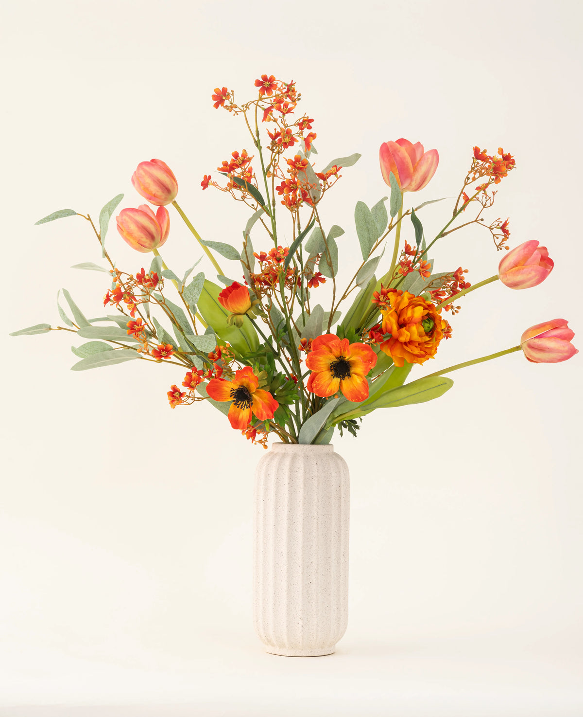Le bouquet de fleurs artificielles en soie Orange Glory, avec des tulipes orange, des coquelicots et des feuilles vertes, est magnifiquement disposé dans un vase blanc texturé sur un fond clair uni.