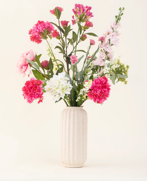 Un vase blanc texturé montre le bouquet de fleurs artificielles en soie Blush Breeze – des fleurs en soie roses, blanches et violet clair avec des feuilles vertes – sur un fond clair uni.
