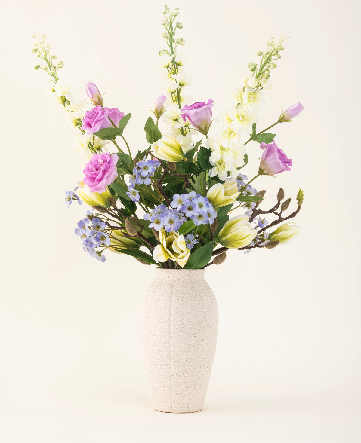 Dans un vase de couleur crème se trouve un bouquet de fleurs artificielles en soie Magnolia Glow avec des fleurs violettes, blanches et jaunes - delphiniums, lisianthus, lys et myosotis sur un fond clair uni.