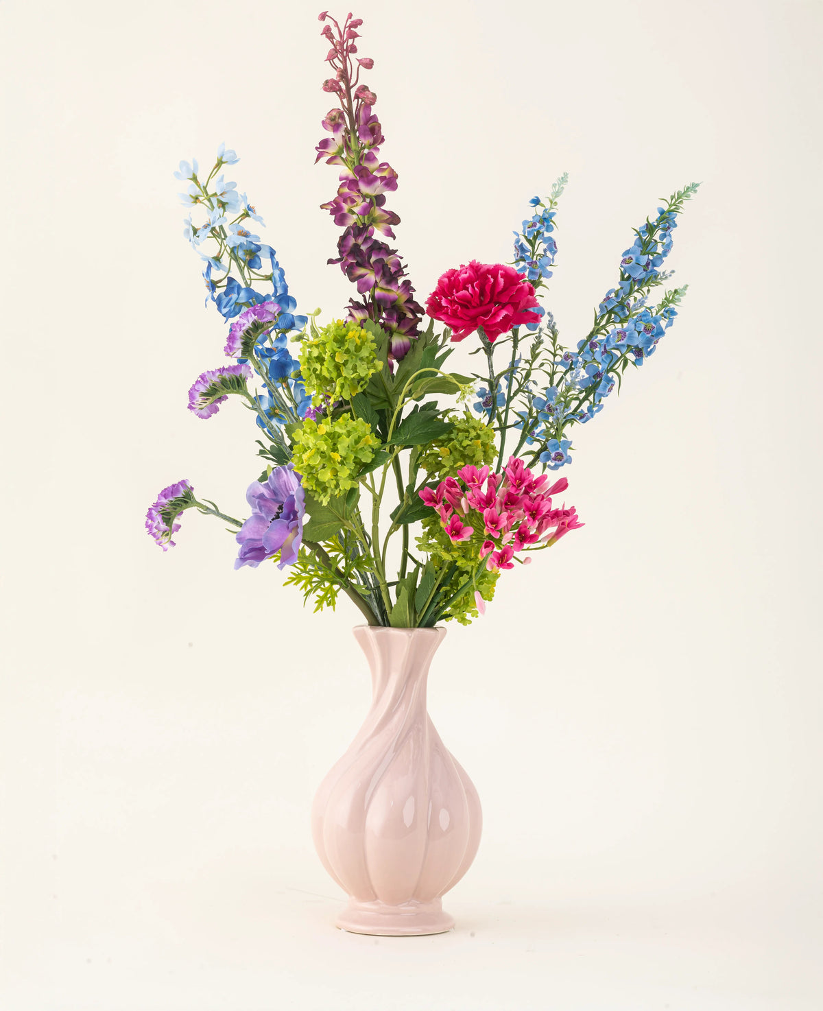 Disposé dans un vase en céramique rose, le bouquet de fleurs artificielles en soie Blossom Breeze présente des fleurs bleues, violettes, rouges et vertes éclatantes – un bouquet floral sans entretien sur un fond clair uni.