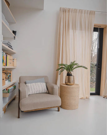 Un coin lecture cosy avec un fauteuil beige, une table d'appoint ronde en osier sur laquelle est posée la plante artificielle Baby Cycas Palm 60 cm, des bibliothèques et des voilages qui pendent au-dessus d'une porte vitrée qui s'ouvre sur le jardin.