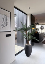 Un intérieur moderne et minimaliste avec un palmier Cycas Classic artificiel de 135 cm dans un pot décoratif élégant près d'une baie vitrée, une œuvre d'art fougère au mur et une cuisine noire élégante avec des lampes suspendues en arrière-plan.