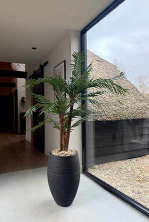 Le palmier artificiel Cycas Classic 135 cm dans un pot décoratif aux lignes épurées est placé près d'une grande fenêtre avec lumière du jour. Dehors, vous pouvez voir un toit de chaume et du gravier. L'intérieur est moderne et minimaliste.