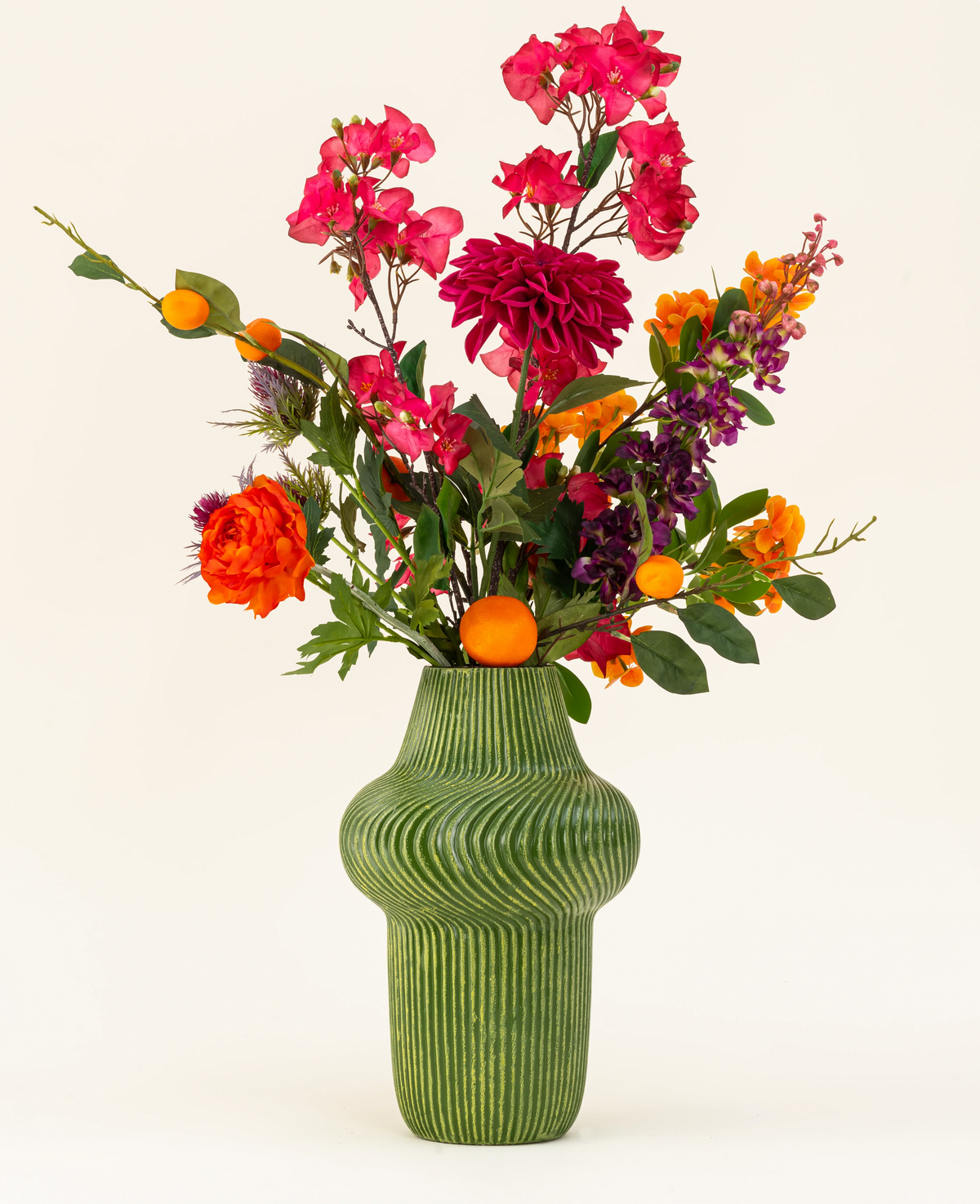 Le bouquet de fleurs artificielles en soie Fiesta Sunset est disposé dans un vase texturé vert et présente des fleurs roses, oranges et violettes avec des feuilles vertes et de petits fruits orange sur un fond clair.