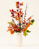 Un vase blanc texturé présente le bouquet de fleurs artificielles en soie Chestnut Charm, composé de feuilles d'automne, de fleurs orange et de feuillage vert - un arrangement artificiel élégant sur un fond clair uni.