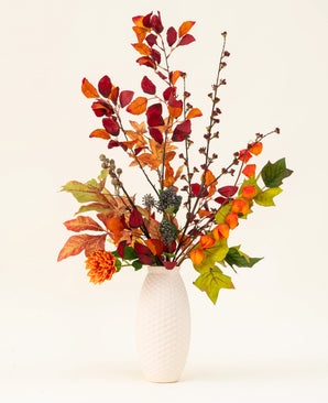 Un vase blanc texturé présente le bouquet de fleurs artificielles en soie Chestnut Charm, composé de feuilles d'automne, de fleurs orange et de feuillage vert - un arrangement artificiel élégant sur un fond clair uni.