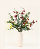 Le bouquet de fleurs artificielles en soie Midwinter Dream contient des feuilles vertes, des branches de pin, des baies rouges et des fleurs blanches dans un vase à structure blanche - parfait pour un bouquet hivernal serein sur un fond clair et uni.