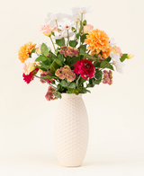 Le bouquet de fleurs artificielles en soie The Wild Bunch, avec des roses rouges, du Dahlia Real Touch, des fleurs roses et des feuilles vertes dans un vase structuré blanc, est présenté sur un fond clair.