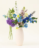 Un vase blanc structuré contient le bouquet de fleurs artificielles en soie Midnight Valley, composé de fleurs Real Touch bleues, violettes et jaunes vibrantes avec plusieurs feuilles vertes sur un fond clair.