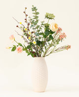 Un vase blanc texturé présente le bouquet de fleurs artificielles en soie Blush Blossom, avec de vraies fleurs – roses rose pâle, fleurs jaunes et feuillage vert – sur un fond clair uni.