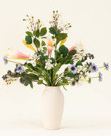 Le bouquet de fleurs artificielles en soie Blush & Bloom – avec des lys roses, des fleurs blanches et violettes et des feuilles vertes luxuriantes – se dresse dans un vase blanc sur un fond clair.