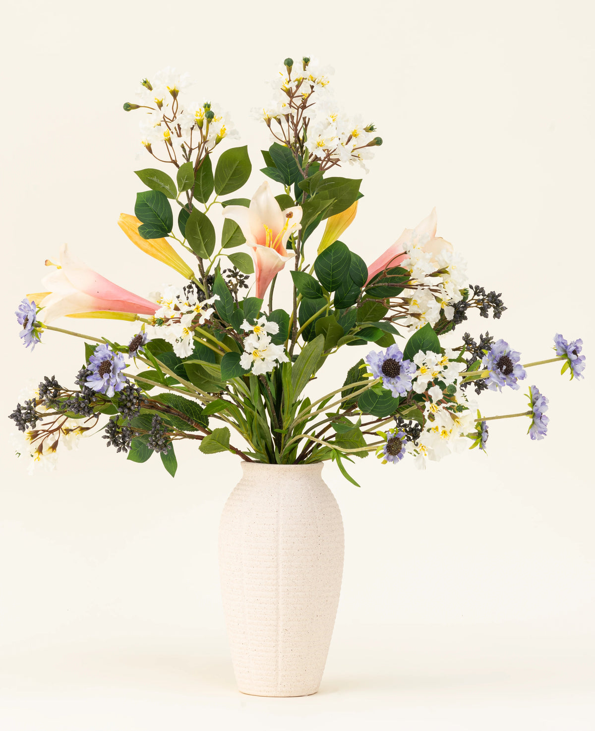 Le bouquet de fleurs artificielles en soie Blush & Bloom – avec des lys roses, des fleurs blanches et violettes et des feuilles vertes luxuriantes – se dresse dans un vase blanc sur un fond clair.