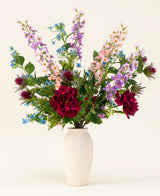 Le bouquet de fleurs artificielles en soie Moonlit Meadow - pivoines rouges, delphiniums roses et violets, fleurs bleues, feuilles vertes et fleurs ressemblant Ă des chardons - se dresse dans un vase Ă structure blanche sur un fond uni et clair.