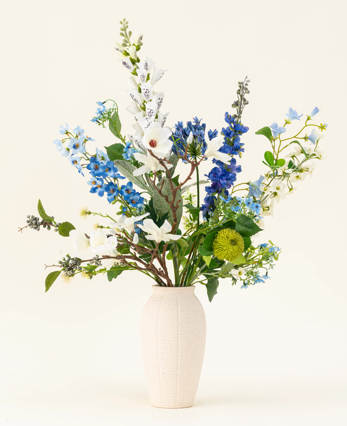 Le bouquet de fleurs artificielles en soie Cool Breeze, avec des lys blancs, des pieds d'alouette bleus et du feuillage vert dans un vase de couleur crème, se détache sur un fond uni pour une touche rafraîchissante.