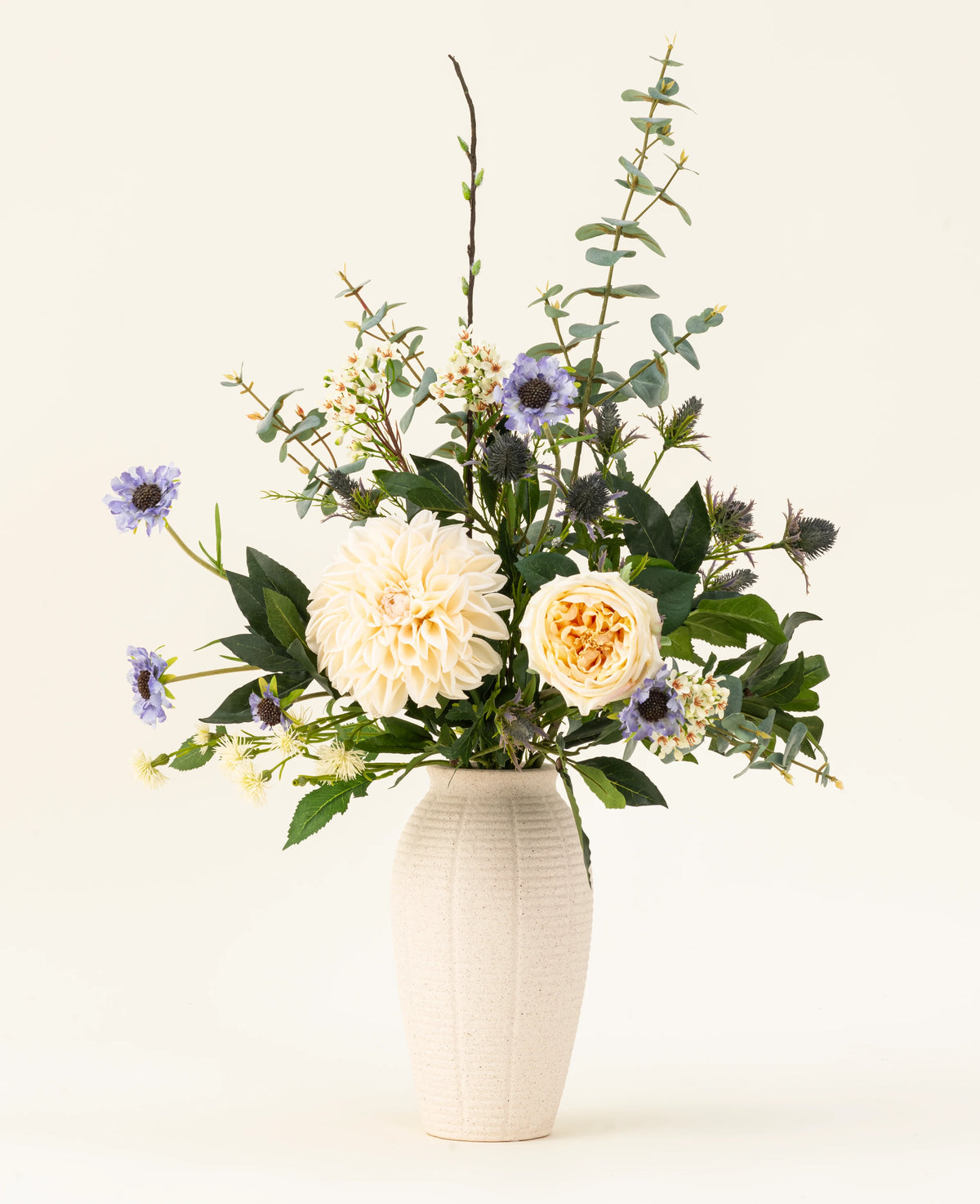 Un vase en céramique blanche contient le bouquet de fleurs artificielles en soie Petal Poetry, avec des dahlias et des roses de couleur crème, des fleurs violet clair, des chardons verts et toutes sortes de verdure sur un fond clair uni.