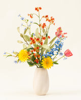 Le bouquet de fleurs artificielles en soie Lemonade Garden comprend des tournesols teintés, des tulipes roses, des delphiniums bleus, des fleurs d'oranger et du feuillage vert dans un vase beige sur un fond clair uni.