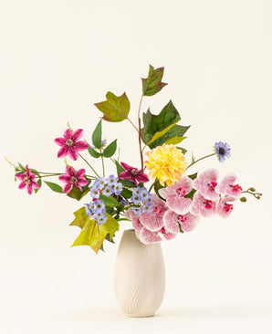 Le bouquet de fleurs artificielles en soie Aurora Garden présente des fleurs au toucher réaliste en rose, jaune, bleu et violet avec des feuilles vertes, disposées dans un vase en céramique blanche sur un fond clair uni.