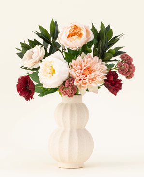 Un vase en céramique de couleur crème avec structure montre le bouquet de fleurs artificielles en soie Jewel Bloom avec des fleurs Real Touch roses, blanches et rouge foncé et des feuilles vertes sur un fond clair uni.