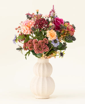 Dans un vase en céramique blanc incurvé se trouve le bouquet de fleurs artificielles en soie Pretty Pastel - un mélange coloré de fleurs durables telles que des roses roses, des dahlias, des fleurs violettes et oranges et de la verdure - magnifiquement disposées sur un fond clair.