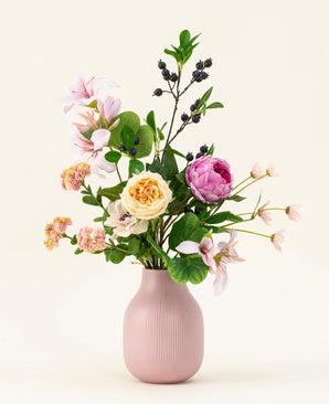 Le bouquet de fleurs artificielles en soie Touch of Cloudlight, présenté dans un vase en céramique rose, contient des pivoines roses, des roses jaunes, de petites fleurs, de la verdure et des branches avec des baies sombres sur un fond clair.
