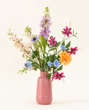 Le bouquet de fleurs artificielles en soie Bloom & Glow présente des fleurs roses, violettes, bleues, orange, blanches et jaunes avec des feuilles vertes dans un vase en céramique rose sur fond clair.