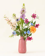 Le bouquet de fleurs artificielles en soie Bloom & Glow présente des fleurs roses, violettes, bleues, orange, blanches et jaunes avec des feuilles vertes dans un vase en céramique rose sur fond clair.