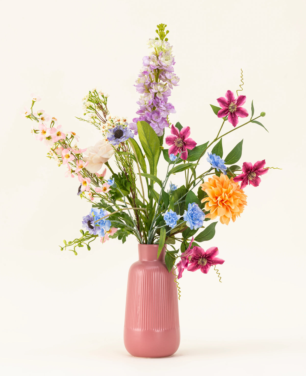 Le bouquet de fleurs artificielles en soie Bloom & Glow présente des fleurs roses, violettes, bleues, orange, blanches et jaunes avec des feuilles vertes dans un vase en céramique rose sur fond clair.