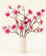 Le bouquet de fleurs artificielles en soie Magnolia rose est disposé dans un vase en céramique blanche ondulée avec des bourgeons verts, créant un affichage élégant sur un fond clair.