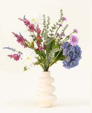 Un vase ondulé de couleur crème contient le bouquet de fleurs artificielles en soie Lavender Kiss, composé de dahlias blancs, de fleurs artificielles lilas, d'hortensias violets, de fleurs roses et violettes et de feuillage vert sur un fond clair uni.