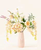 Un vase en céramique rose pastel présente le bouquet de fleurs artificielles en soie Blushing Beauty, composé d'hortensias aux couleurs douces, de roses et de verdure assortie, magnifiquement disposés sur un fond neutre pâle.