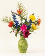 Un vase vert moucheté montre le bouquet de fleurs artificielles en soie Hibiscus Fiesta - avec des fleurs roses, rouges et jaunes, des delphiniums bleus, des oiseaux de paradis orange et des feuilles de palmier vertes - sur un fond clair uni.