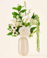Dans un vase en céramique blanche se trouve le bouquet « Green Elegance » de fleurs artificielles en soie, un arrangement d'hortensias blancs, de callas, d'anémones, de feuilles vertes et de verdure suspendue sur un fond clair uni.