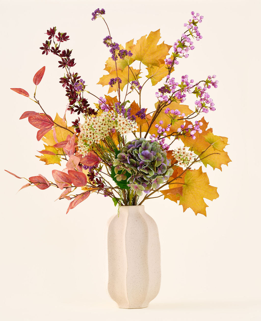 Le bouquet de fleurs artificielles en soie 'Wild Amethyst', avec des feuilles d'automne, des fleurs violettes et blanches et des hortensias verts, est présenté dans un vase en céramique blanche sur un fond uni et clair.