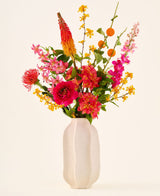 Dans un vase en céramique blanche se trouve le bouquet de fleurs artificielles en soie « Summer Blush », composé de fleurs roses, jaunes et orange avec des feuilles vertes, sur un fond clair uni.