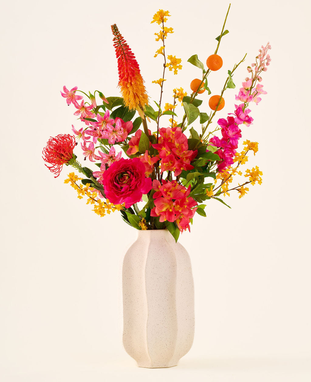 Dans un vase en céramique blanche se trouve le bouquet de fleurs artificielles en soie « Summer Blush », composé de fleurs roses, jaunes et orange avec des feuilles vertes, sur un fond clair uni.