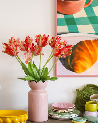 Sur une table avec de la vaisselle colorée se trouve un vase rose contenant la fleur artificielle tulipe de perruche française de 76 cm rouge/jaune, tandis qu'en arrière-plan on peut voir en partie une peinture encadrée d'un croissant et d'une tasse.