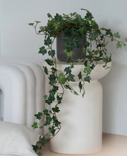 Un lierre artificiel Deluxe Green, un lierre suspendu de 90 cm dans un pot gris, se dresse sur une base sculpturale blanche et moderne à côté d'une couverture blanche pliée et d'un coussin blanc structuré.