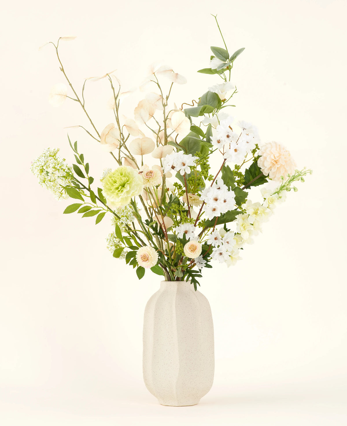 Le bouquet de fleurs artificielles en soie Claire de Lune présente de délicates fleurs blanches, jaune pâle et vertes dans un vase en céramique blanche, accentuées par une verdure luxuriante sur un fond clair.