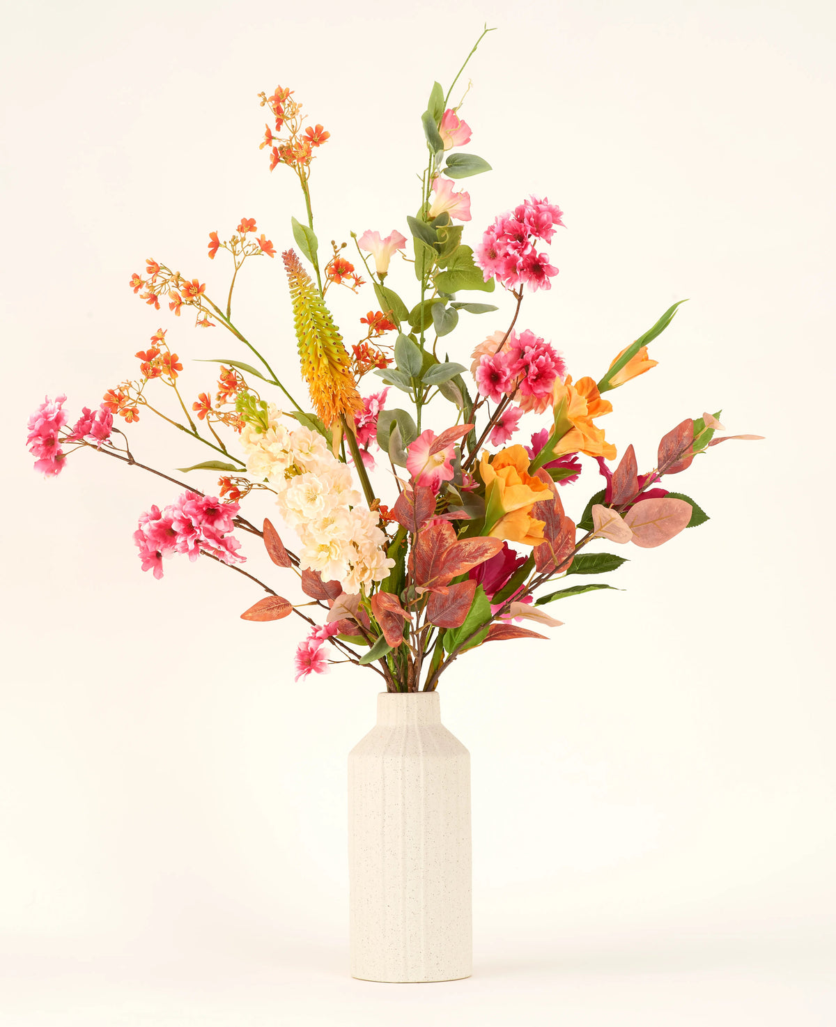 Dans un vase à structure blanche se trouve le bouquet de fleurs artificielles en soie Wild Peach avec des fleurs artificielles colorées - roses, orange, jaunes et blanches - et des feuilles vertes et rougeâtres sur un fond clair uni.