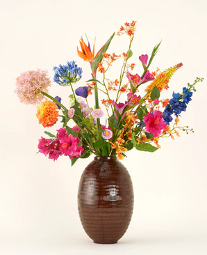 Dans un vase à structure marron se trouve le bouquet de fleurs artificielles en soie Tropical Firework - un arrangement de fleurs en soie orange, roses, violettes, jaunes et bleues avec des feuilles vertes sur un fond clair uni.